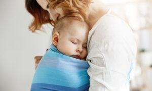 Precious little newborn boy having deep sleep at day on mother chest in blue baby sling. mom kissing baby head and feeling relaxed and delight. family concept.